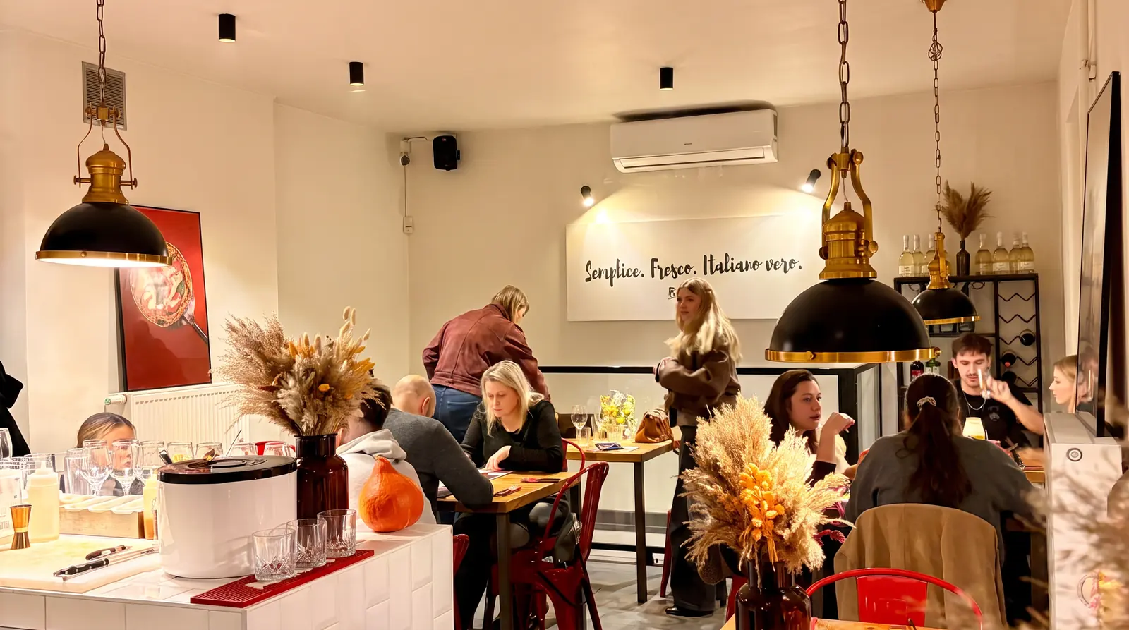 Sala Bottegas Saska Kępa przy ul. Francuskiej 24 wieczorem — mosiężne lampy nad drewnianymi stołami, czerwone krzesła, suszone kwiaty i napis Semplice. Fresco. Italiano vero.